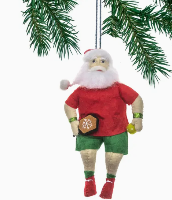 A handcrafted felt ornament resembling Santa Claus wearing a red shirt and green shorts, with a plush green Christmas tree in the background.