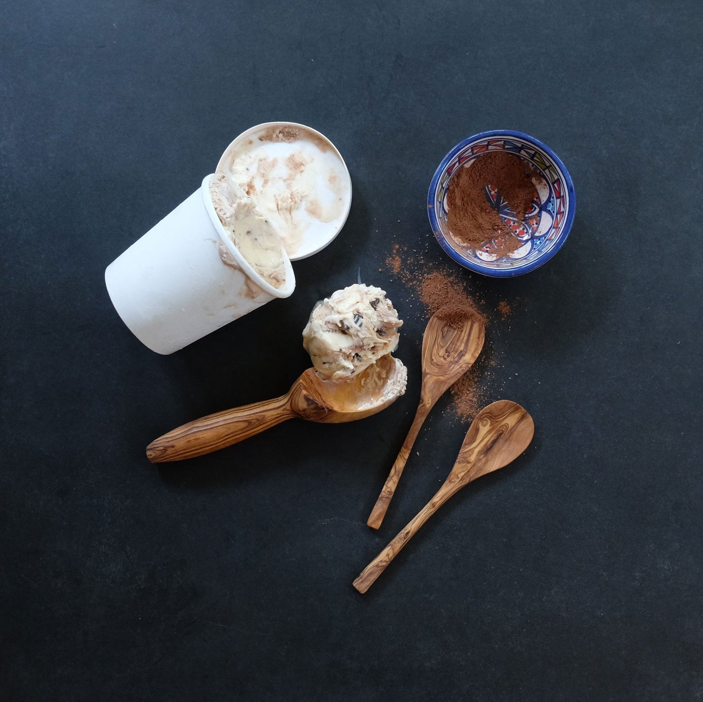 Ice cream scoop with ice cream, a container of ice cream, and a small bowl on a dark surface.