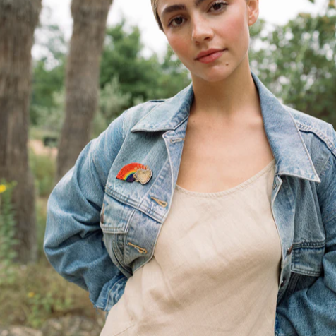 A person wearing a denim jacket and a beaded rainbow brooch pin on the lapel.