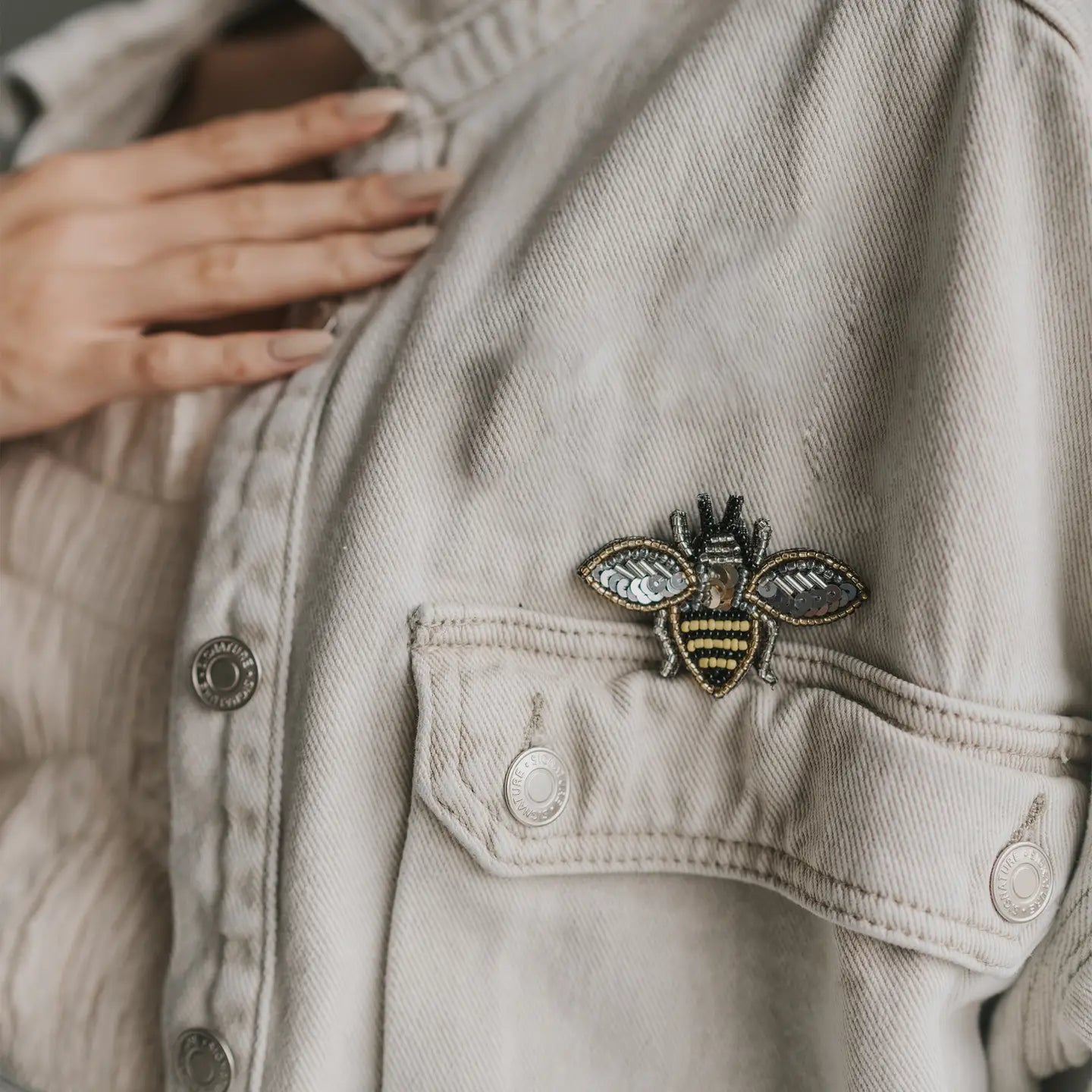 Beige denim jacket with a decorative bee pin on the chest pocket.