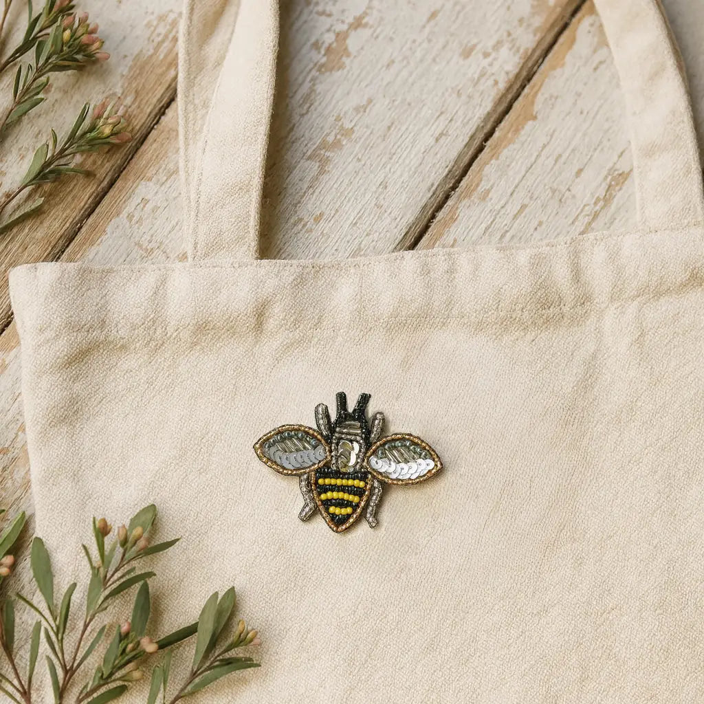 Beige tote bag with a bee brooch pin on a wooden surface with greenery.