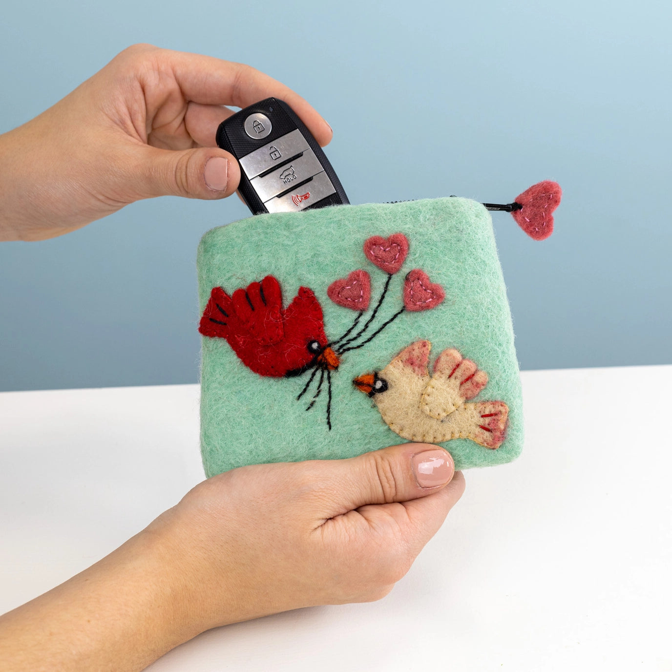 Hand holding a car key over a felt pouch with embroidered designs on a light blue background