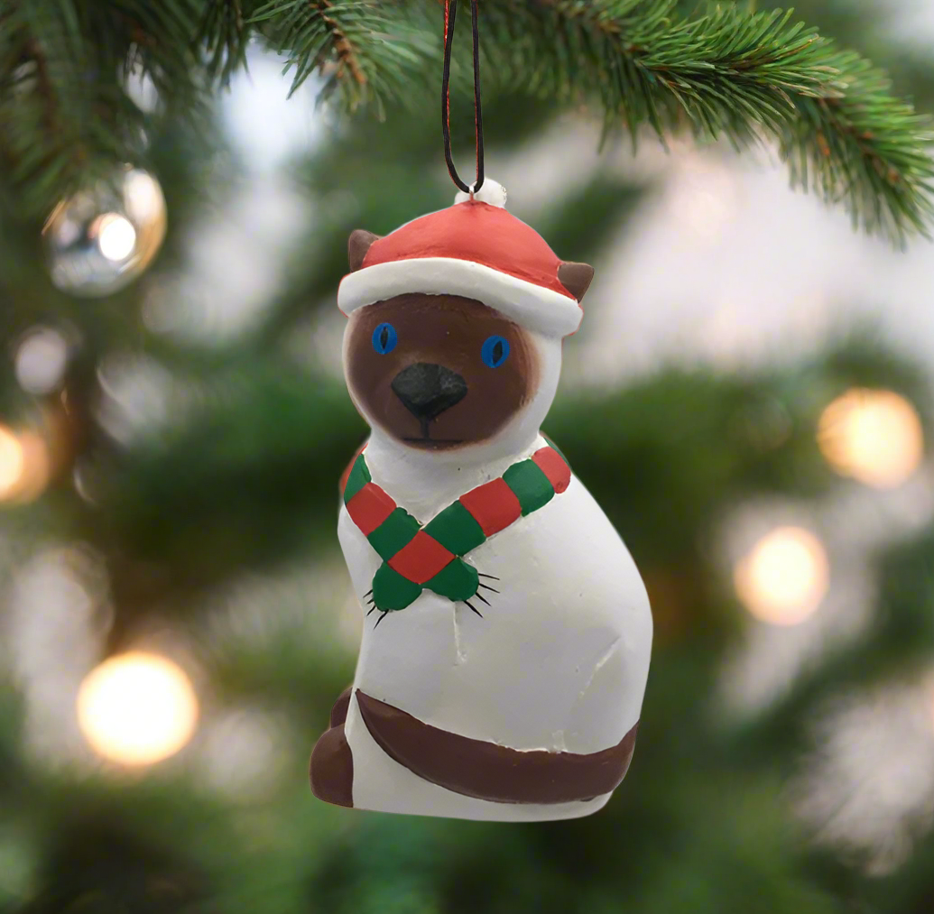 A hand-carved balsa wood ornament depicting a Siamese cat wearing a festive hat and scarf, painted in multiple colors.