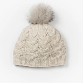 A beige wool beanie with a cable knit design and a gray faux fur pompom on top.
