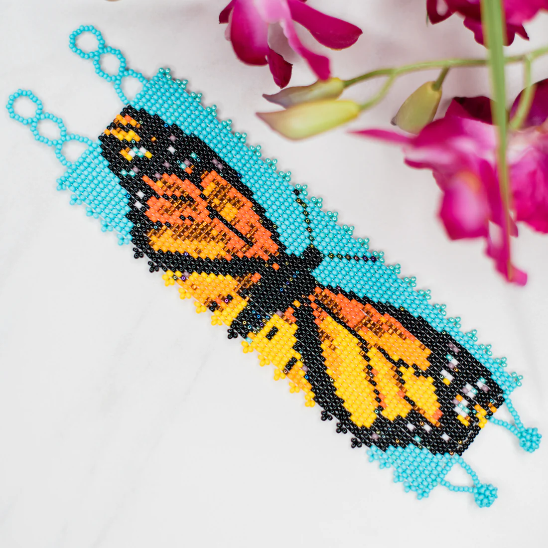 Beaded butterfly bracelet with blue, orange, and black colors on a white background with pink flowers.