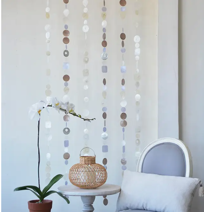 A decorative capiz shell strand hanging vertically on a wall, featuring circular and square shaped shells in beige, white, and blue colors, with a metal ring at the top for hanging. There is a small round table below with a woven basket and a potted plant on it, and a white cushioned chair to the side.