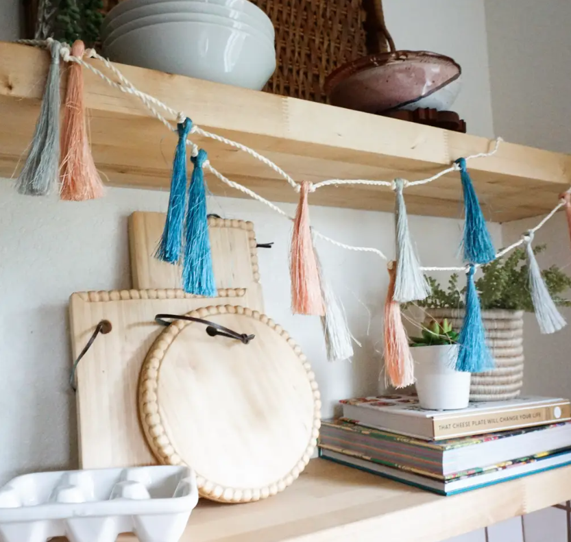 Decorative tassel garland on a wooden shelf with books and plants.
