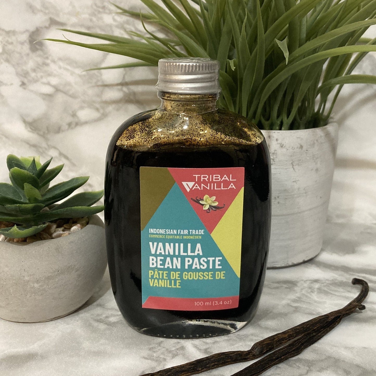 A 100mL glass bottle of Tribal Vanilla Bean Paste with a colorful label, placed on a marble surface with a cactus plant and a piece of vanilla bean pod in the background.